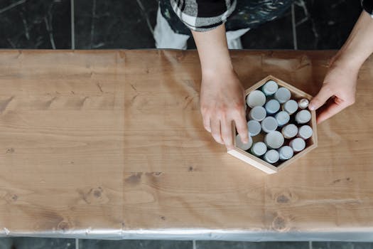 An artist's hands arranging paint bottles in a hexagonal box on a work table.