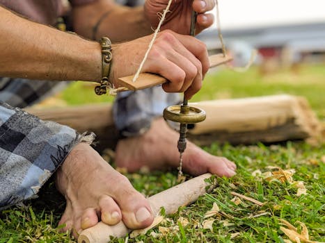 Close-up of a man's hands and feet working with traditional woodworking tools outdoors on grass.