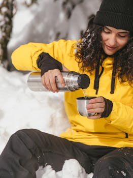 Woman in yellow jacket pouring hot drink from thermos in snowy outdoor setting.